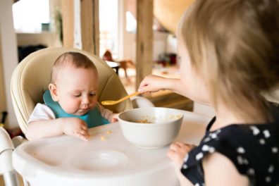 Étvágytalan baba, szőke kislány etet kiskanállal, egy kisfiú babát, aki etetőszékben ül, kép.
