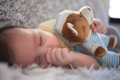 fejlődés elősegítése az első félévben, barna hajú csecsemő oldalt feküdve, hüvelykujja a szájában, macit ölel magához és alszik, kép.