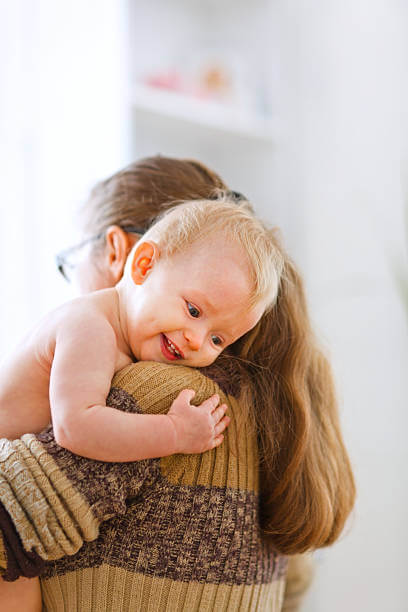 Egészséges csecsemő fejlődése, hosszú barna hajú anyuka vállát átölelő meztelen, szőke kisbaba, kép.
