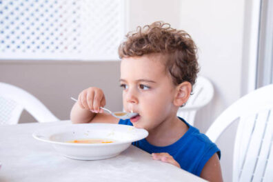 Önálló evésre szoktatás, kék ujjatlan pólóban egy barna hajú kisfiú, tányérból, kanállal eszik az asztalnál, kép.